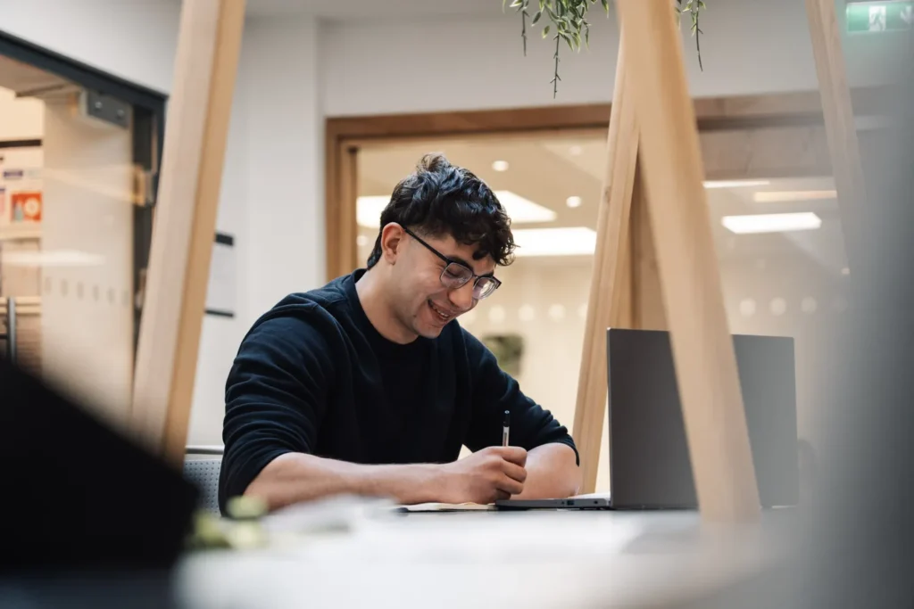 male student sat at desk with a laptop writing with a pen